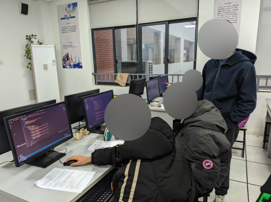 Three students in a computer classroom, with two students working on their respective computers and one student standing behind them.